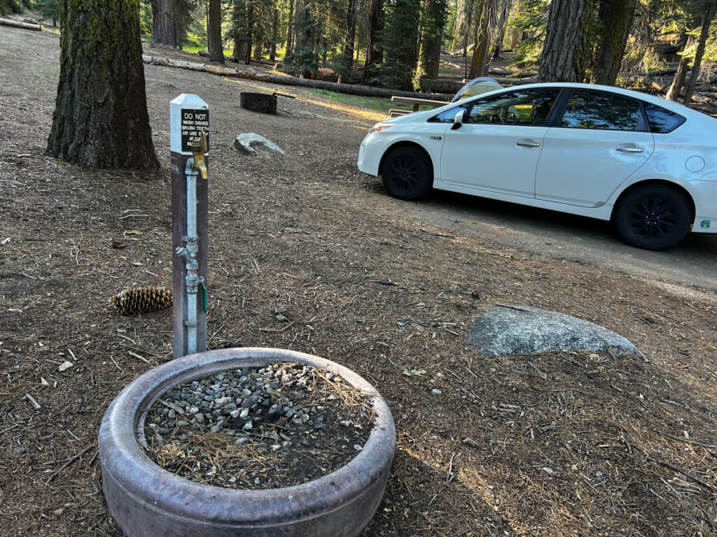 Image of a campsite with a water spigot in the foreground, a white Prius in the background