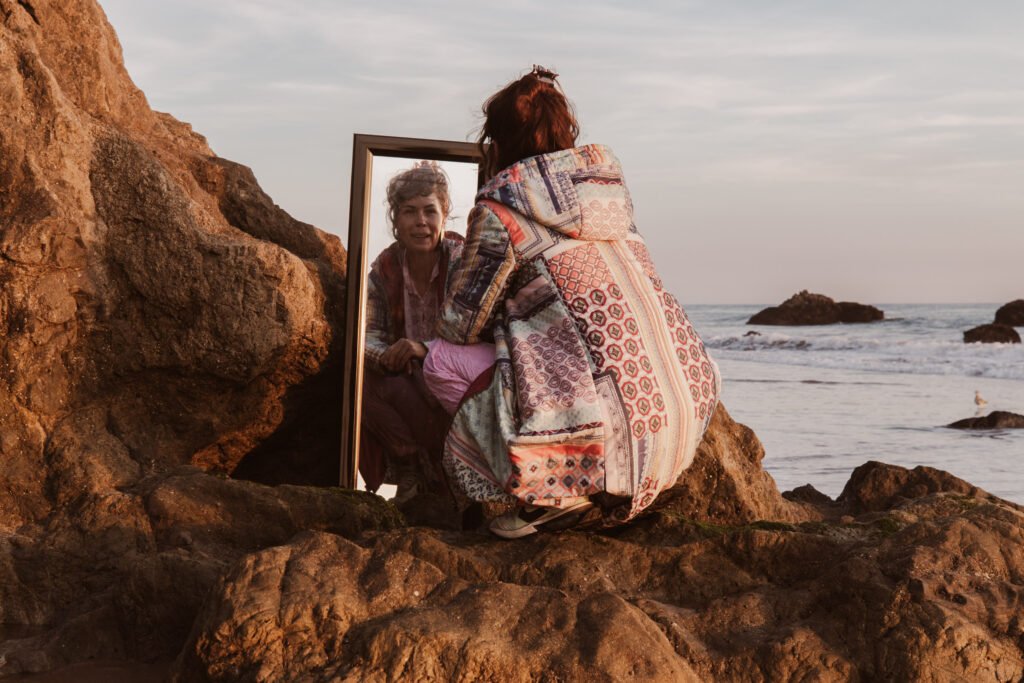 A model crouches on a rock at the beach, wearing a quilted jacket. She's looking at the camera through a mirror.