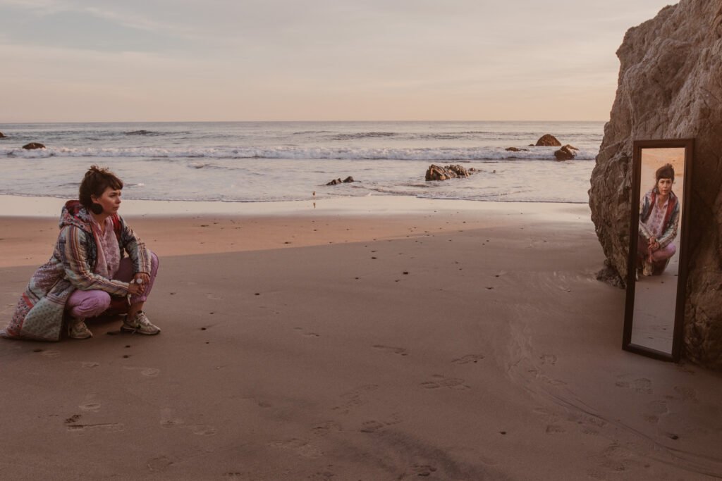 A model crouches on the beach, wearing a quilted jacket. She's looking at the camera through a mirror.