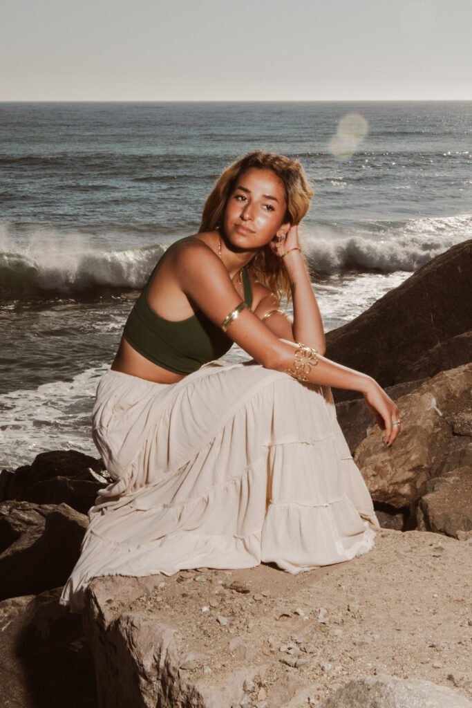 A model crouches on some rocks in front of the ocean.