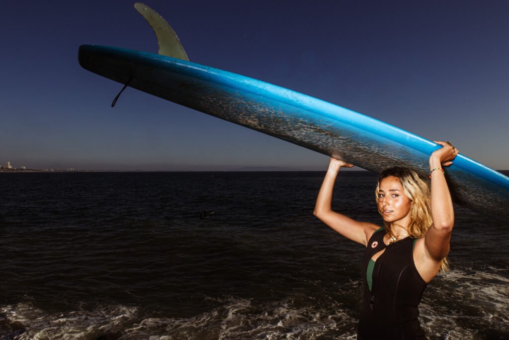 A surfer with bleached blonde hair holds a blue surfboard over her head.