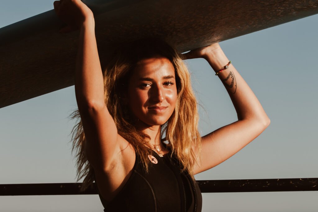 A surfer with bleached blonde hair holds a surfboard over her head.