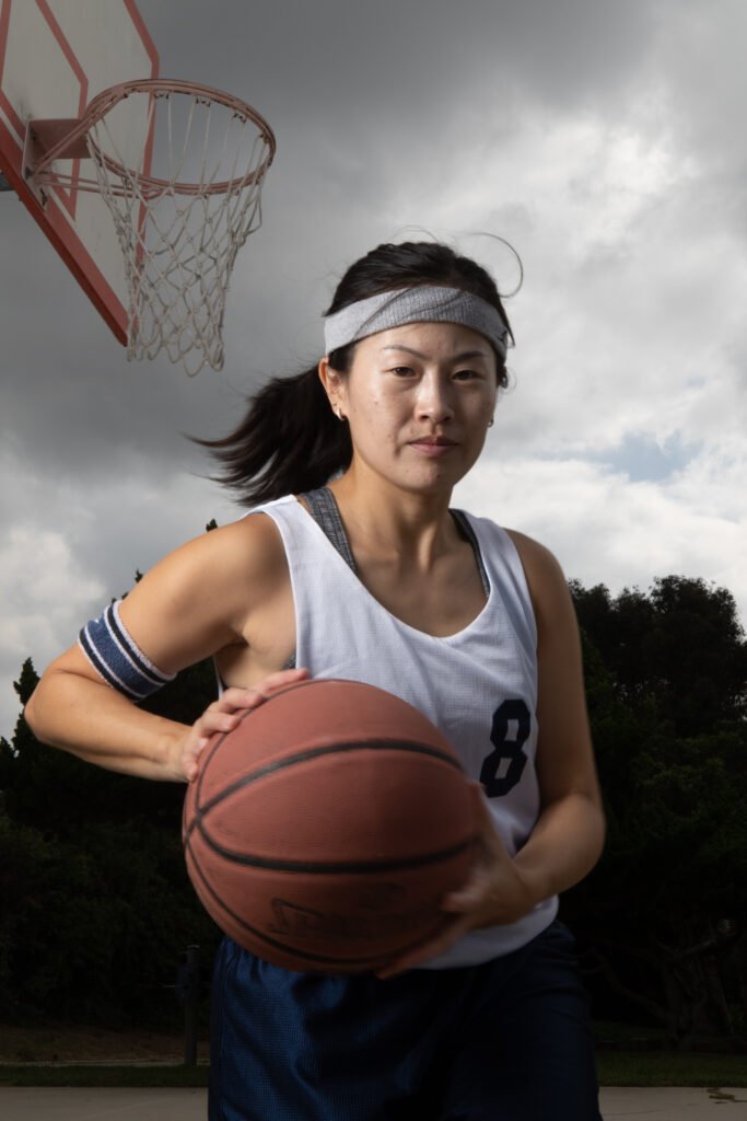 A woman holds a basketball under an outdoor hoop, looking as if she is just about to dribble the ball.